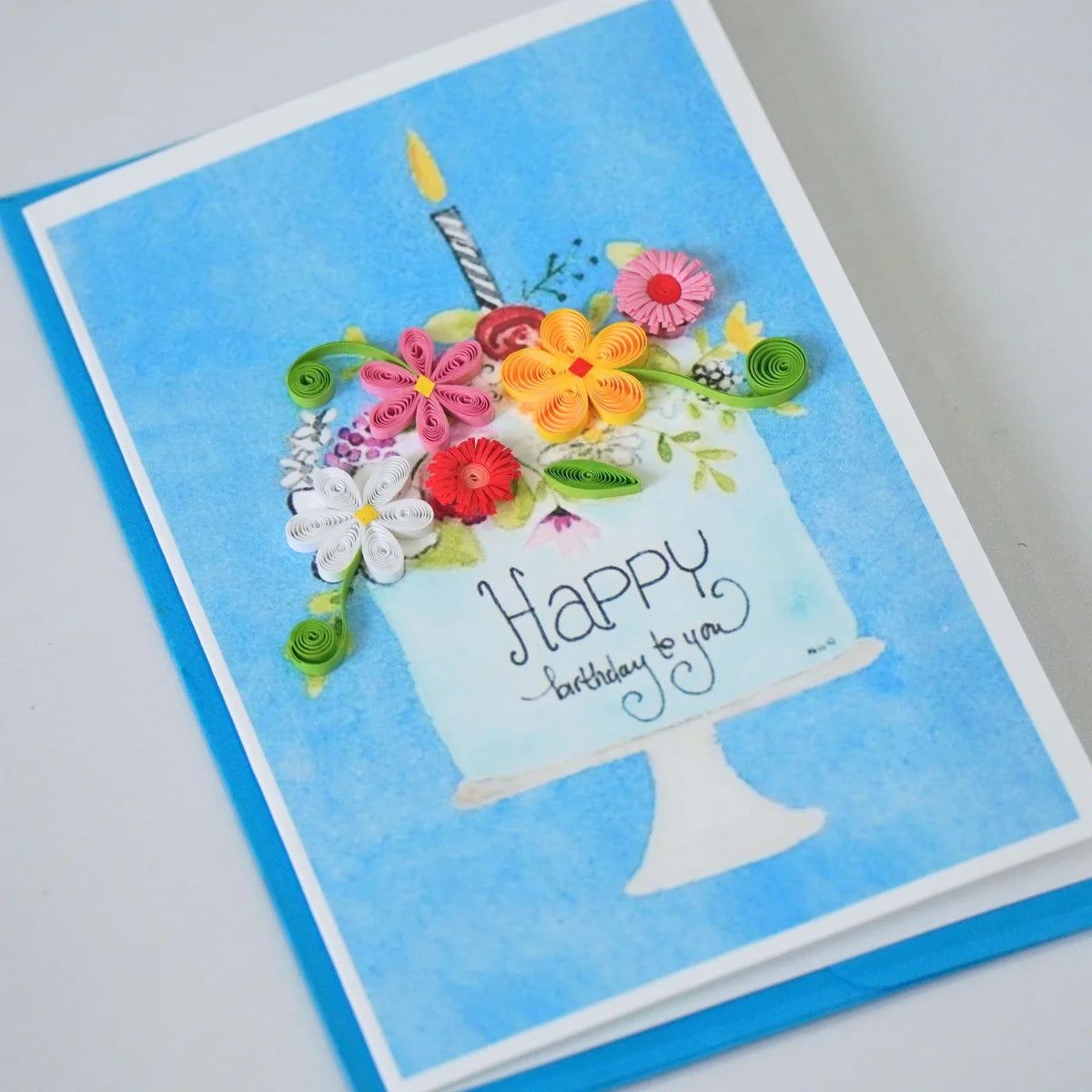 Angled view of happy Birthday Cake quilled greeting card with colorful paper flowers and a candle