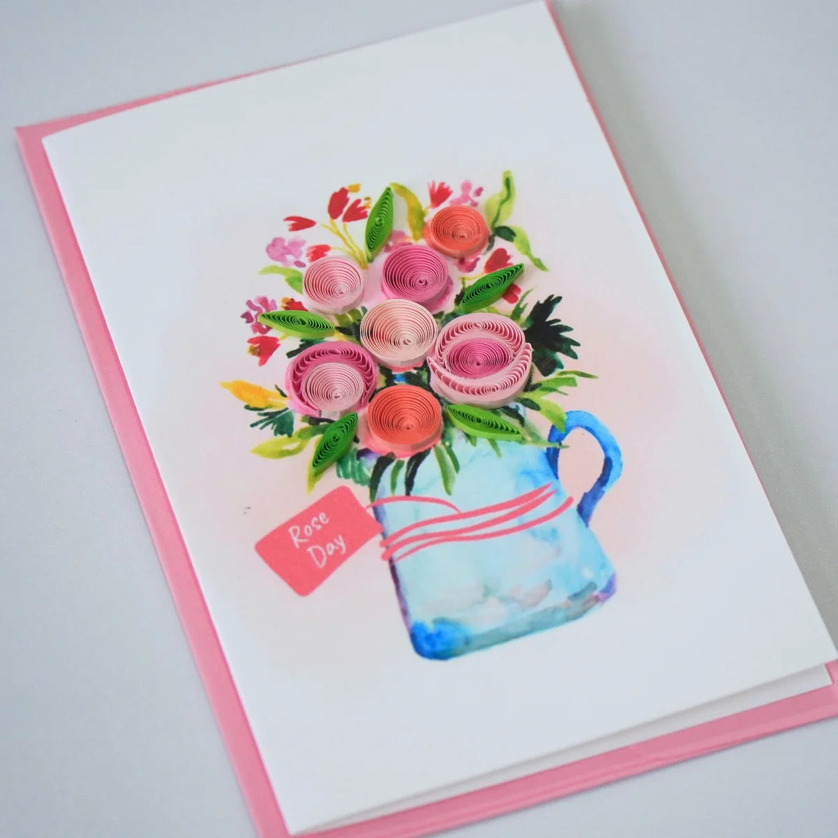 Angled view of rose Day quilled greeting card with pink paper roses in an illustrated blue jug