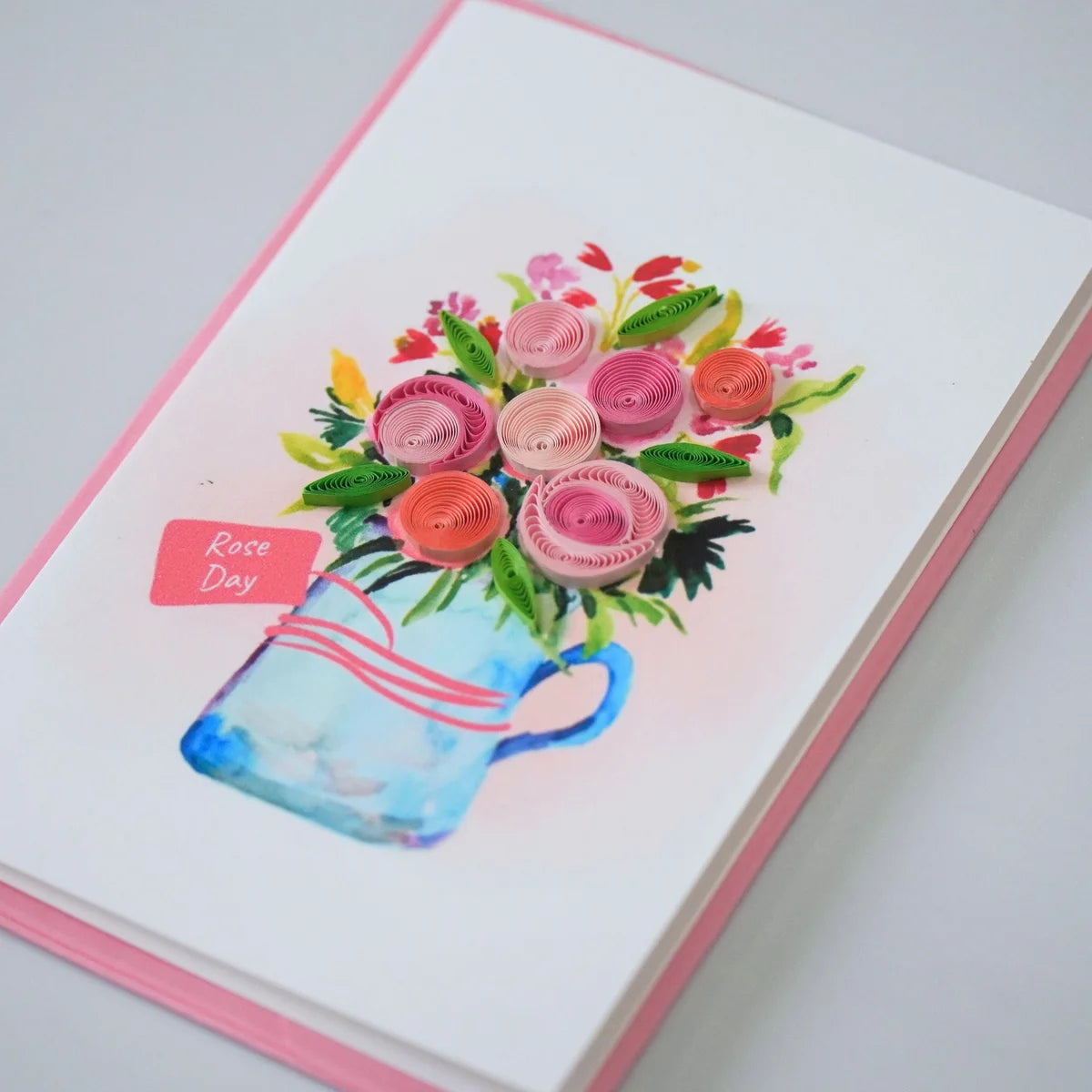 Side view showing pink and peach coiled paper roses above a blue jug and Rose Day tag